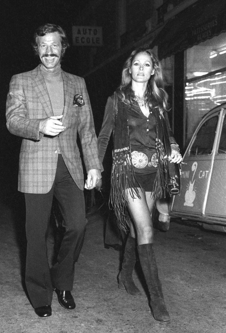 Jean-Paul Belmondo smiling while wearing a checkered coat, turtle-neck shirt, and pants with his wife Ursula Andress in Paris